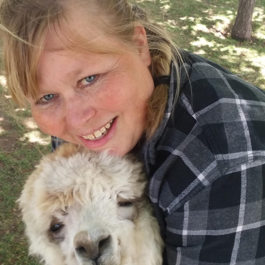A smiling woman with blonde hair wearing a flannel shirt leans down to hug a fluffy white alpaca