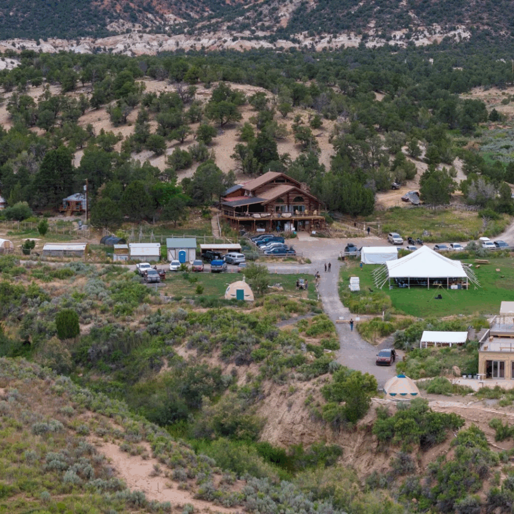 A rustic guest ranch nestled in a high desert valley of southern Utah, featuring a large lodge building, cabins, yurts, and an event tent surrounded by green trees and rugged hills